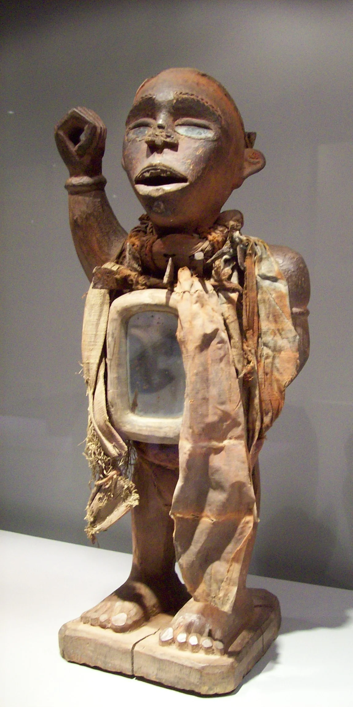 Magische Figur (Bakongo, Mayombe, Dem. Republik Kongo)
Die Figur weist sowohl ein Spiegelkästechen (nur an der Brustseite!) als auch einige eingeschlagene Nägel und Klingen auf. Das Gesicht ist fein ausgearbeitet, ein Fischgerätemuster betont die plastisch erhöhten Augenbrauen, die Lippen des leicht geöffneten Mundes sind sorgfältig geformt; die obere Zahnreihe zeigt die für die Yombe charakteristische Lückenaussplitterung. Die Kopfbedeckung (mit Zauberbehälter) fehlt.